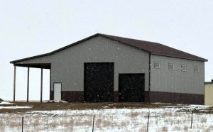 A grey metal barn with a brown roof sits in a snowy landscape. It has two large dark doors, a smaller white door, and four square windows.