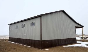 A grey metal building with brown trim and four windows sits on dirt ground with patches of snow.
