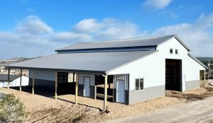 A large, modern metal barn with gray siding and a gabled roof stands on a dirt lot under a partly cloudy sky.