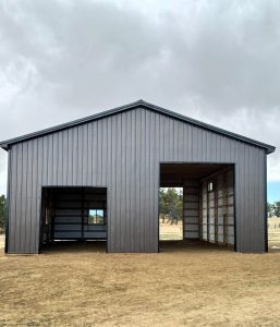 A gray metal building with large open doorways stands on a dirt lot under a cloudy sky.