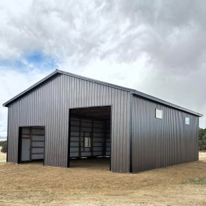 A dark metal barn with a large open entrance, a side door, and two windows under a cloudy sky.