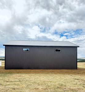 A dark brown, metal-sided building with a metal roof sits on a dry, grassy field under a cloudy sky.