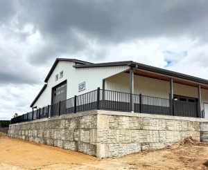 A modern metal building with large garage doors sits on a raised stone foundation, surrounded by a black metal fence under a cloudy sky.