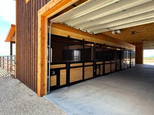 Open garage with wooden and metal horse stalls inside a barn, showing concrete flooring and partially opened overhead doors.