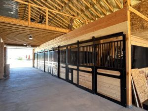A wooden horse stable with black metal railings inside a spacious barn with an exposed beam ceiling and concrete floor.