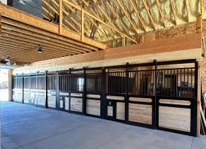 A row of empty horse stalls with black metal and wood panels inside a barn with exposed beams and insulated ceiling.