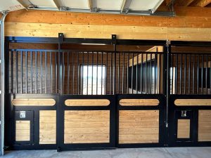 A wooden and metal horse stall with a sliding door and barred upper panels inside a barn. The interior features natural wood walls and a window.