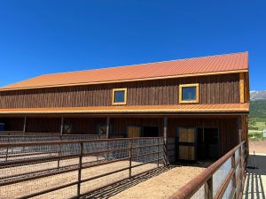 A wooden barn with a metal roof and fenced enclosures stands under a clear blue sky with mountains visible in the background.