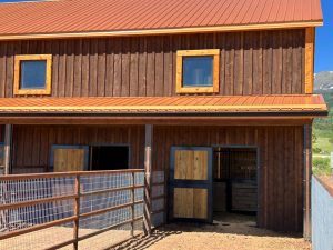 A wooden barn with a metal roof, two square windows on the upper level, and open double doors on the lower level, next to a metal fence.