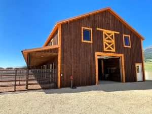 A large wooden barn with a gravel driveway, open garage door, covered side area, and fenced enclosure, set against a blue sky and grassy landscape.