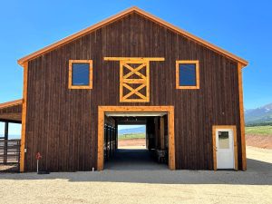 A large wooden barn with two square windows, a central open garage door, and a white side door, set against a clear blue sky and open fields.