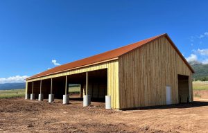 A large wooden barn with a red metal roof and open bays stands on a dirt lot under a clear blue sky.