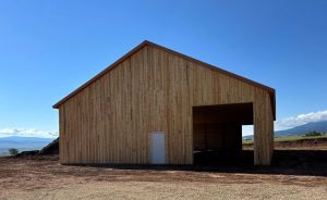 A large wooden barn with a pitched roof and a wide open entry, situated on bare ground with mountains visible in the background under a clear sky.