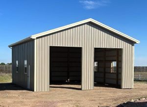 A beige metal garage or storage building with two large open doors, standing on a dirt lot under a clear blue sky.