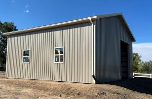 A beige metal building with three windows and a large open garage door stands on bare ground under a clear, blue sky.