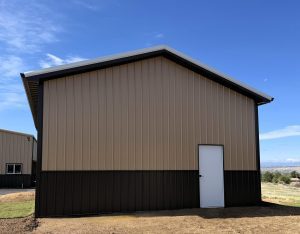 A tan and dark brown metal-sided building with a white door stands on a dirt and grass lot under a clear blue sky.