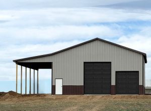 A metal barn with a covered side, a white door, and two dark garage doors under a cloudy sky.