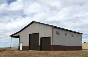 A gray and brown metal barn with multiple garage doors and windows sits under a cloudy sky.