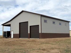 A light gray metal building with a brown trim and roof has two large garage doors, a smaller door, an open bay with a trailer, and four windows.