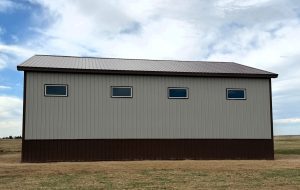 A long, gray metal building with a brown roof and trim, featuring four rectangular windows under a cloudy sky.