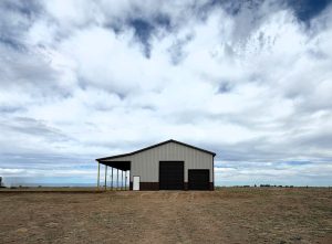 A grey metal building with a covered porch and two garage doors sits under a cloudy sky in an open field.