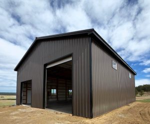 A brown metal barn with open garage doors under a cloudy blue sky.