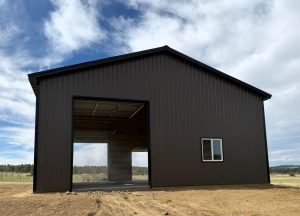 A dark brown, corrugated metal building with a large open doorway and a single window stands under a cloudy sky.
