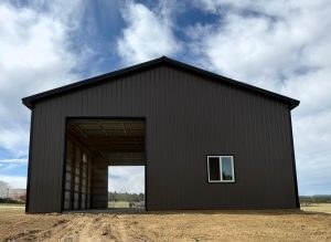 A dark brown metal building with an open bay door and one window under a cloudy sky.