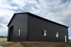A dark metal barn with three windows and a large open doorway under a cloudy sky.
