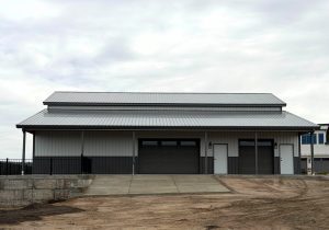 A large, modern metal building with three garage doors and a sloped metal roof, set on a concrete driveway with bare earth in the foreground.