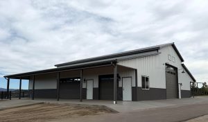 A large, modern metal building with multiple garage doors, windows, and a covered porch area, set on a concrete lot under a cloudy sky.