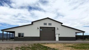 Large metal building with a high central garage door, flanked by two smaller sections, outdoor lights, and a partially cloudy sky in the background.