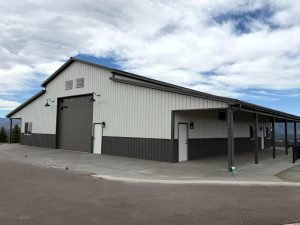 A large gray and white metal building with a tall garage door, small windows, and a covered side walkway, set on a paved surface under a partly cloudy sky.