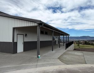 A modern metal building with a covered concrete walkway and railings overlooks a scenic landscape with hills and partly cloudy skies.