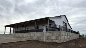 A large, modern metal building with a sloped roof sits on a raised stone foundation, surrounded by a black fence and a concrete driveway under a cloudy sky.