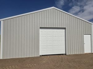 A light gray metal building with a white garage door and a white entry door, set on bare dirt under a partly cloudy sky.