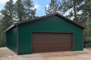 A green metal garage with a large brown roll-up door stands on a dirt lot, surrounded by tall pine trees under a partly cloudy sky.