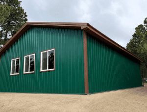 A green metal building with a brown roof trim and three windows on one side, surrounded by trees and a gravel area.