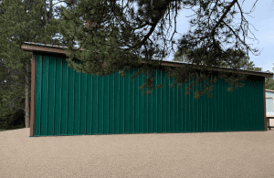 A green metal-sided building with a brown roof stands on a gravel surface, partially shaded by pine tree branches in the foreground.