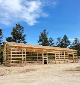 A wooden frame structure under construction stands on a dirt lot, surrounded by trees and under a partly cloudy blue sky.