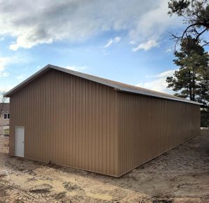 A large, tan metal building with a sloped roof and a single white door sits on bare ground under a partly cloudy sky.