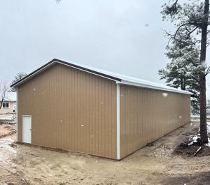 A tan metal storage building with a white door sits on a dirt lot, surrounded by trees and light snowfall under a cloudy sky.