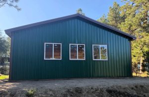 Green metal-sided building with three rectangular windows, situated on bare ground, surrounded by trees under a clear sky.