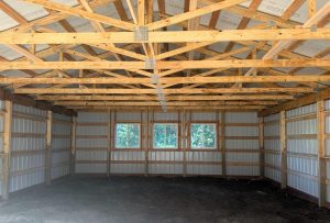 Interior of an unfinished metal-sided building with exposed wooden trusses, three windows, and a dirt floor.