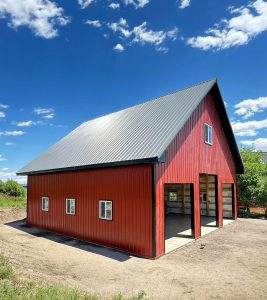 A red barn with a steep metal roof and multiple windows stands on a dirt area under a blue sky with scattered clouds.
