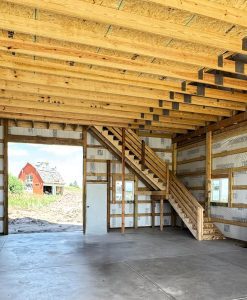 Interior view of a partially constructed building with exposed wooden beams, insulation, concrete floor, and a wooden staircase; red barn visible outside through the open doorway.