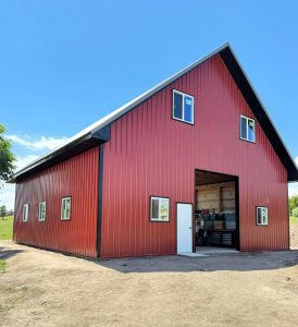 A large red metal barn with black trim, several windows, a white door, and an open garage-style entrance under a clear blue sky.