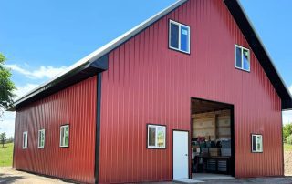 A large red metal barn with black trim, several windows, a white door, and an open garage-style entrance under a clear blue sky.