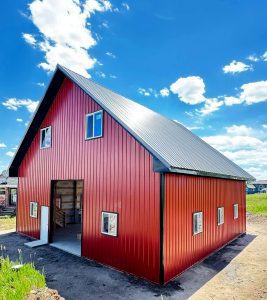 A red metal barn with a steep black roof, multiple windows, and an open doorway stands under a blue sky with scattered clouds.