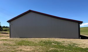 A large gray metal building with a red roof stands on a grassy area under a clear blue sky.